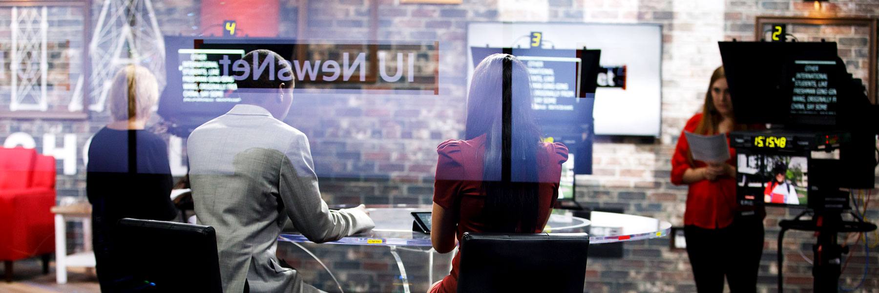 One student operates a teleprompter as two other students report news in a video production studio.