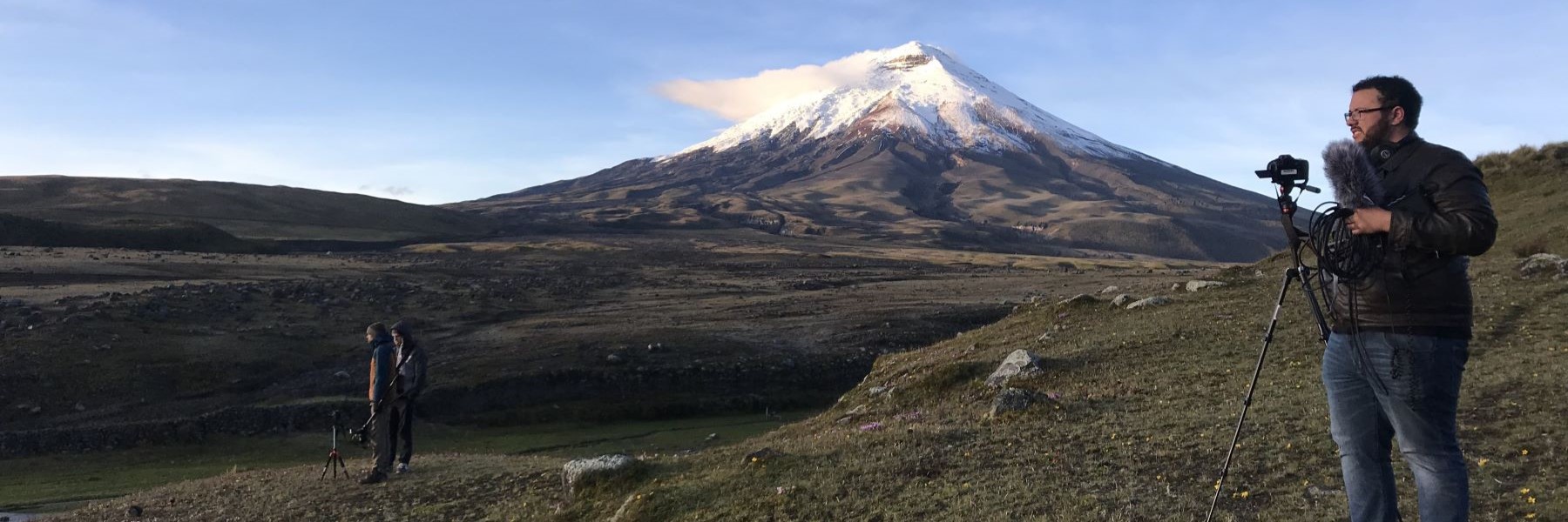 Person adjusting a video camera on a tripod while standing out in a vast field with a snow-capped mountain in the background.