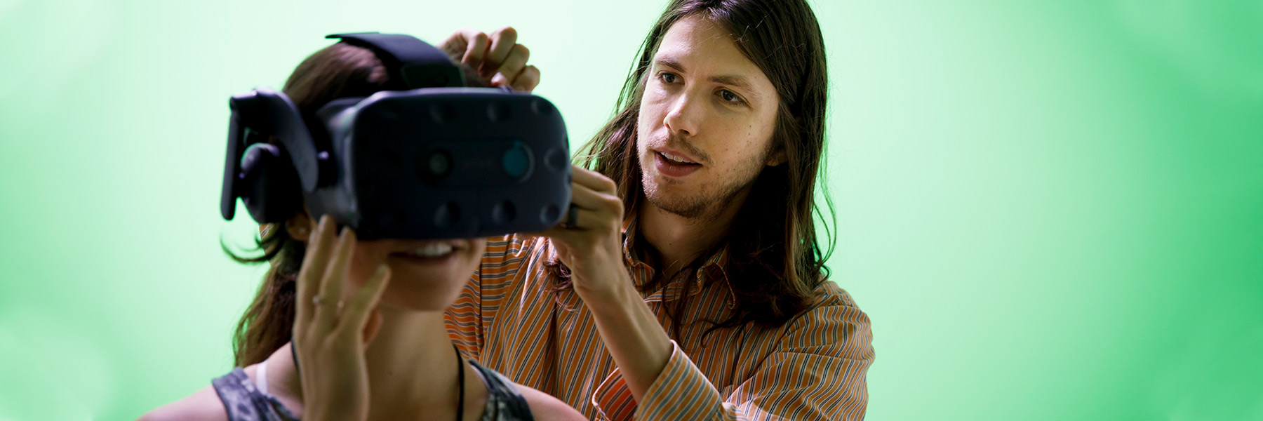 A student helps another student put on a virtual reality headset in front of a green screen.