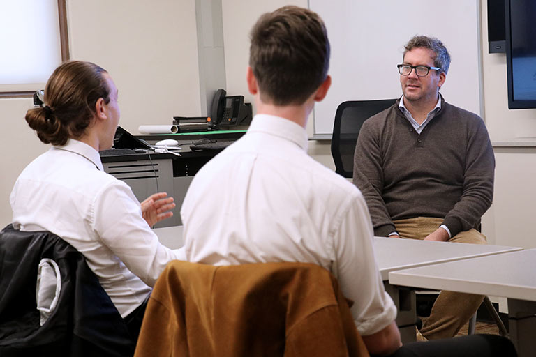 Two students sit across a table from a man during an interview.